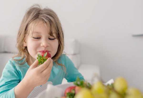 Девочка ест клубнику. На столе виднеются фрукты
