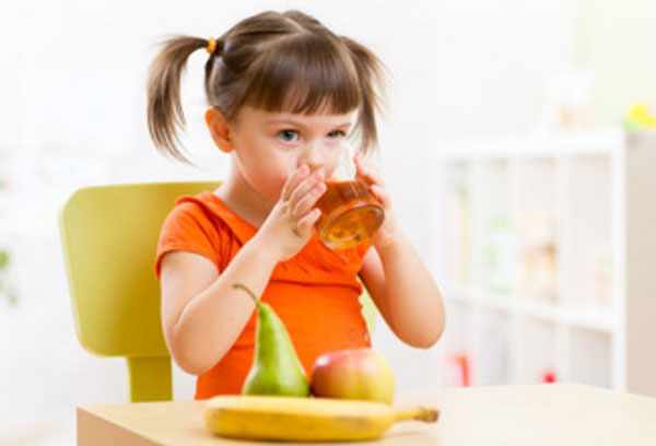 Девочка пьет яблочный сок. На столе лежит банан, груша и яблоко