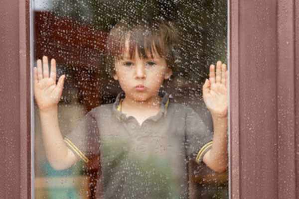 Грустный мальчик стоит у окна, за которым идет дождь