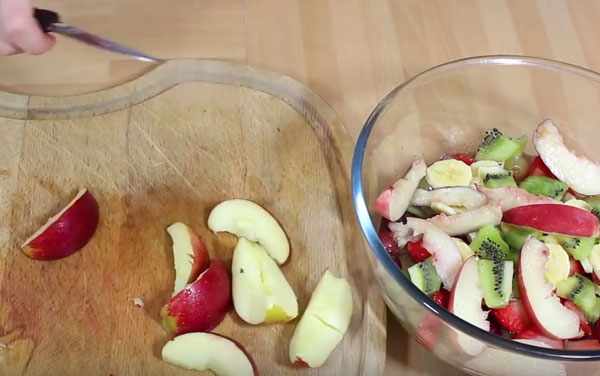 Яблоко нарезано на дольки, лежит на достояке. Рядом мисочка с остальными ингредиентами салата