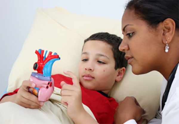 Ребенок лежит в постели и держит модель сердца, рядом доктор