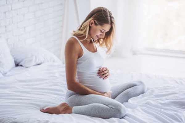 Беременная женщина сидит на кровати, гладит свой живот и улыбается
