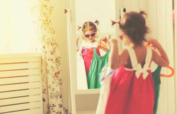 Маленькая девочка в солнцезащитных очках стоит перед зеркалом, держа в руках вешалки с платьями