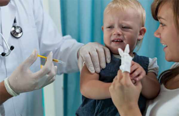 Маленькому ребенку делают прививку в плечо. Он сидит у мамы на руках.