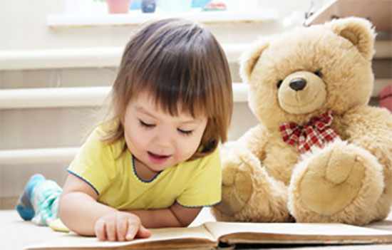 Маленькая девочка читает книгу лежа на полу, рядом сидит плюшевый медведь